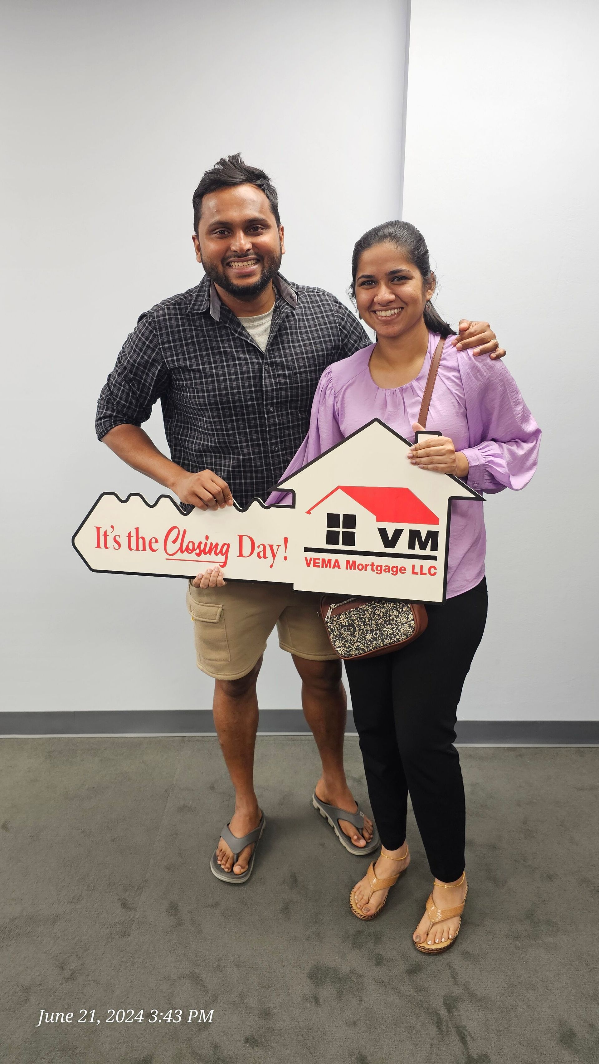 Two smiling people holding a house-shaped sign and a plaque, standing indoors against a plain wall.