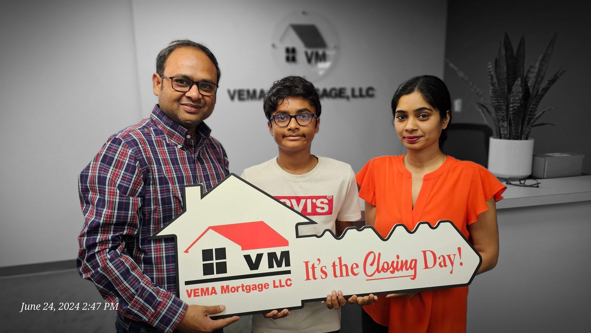 Three people hold a VM logo sign reading “It’s the Closing Day!” in an office setting