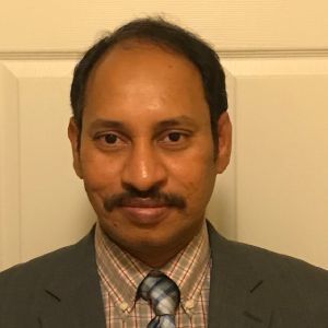 Man in a suit and tie posing against a beige door, facing the camera with a slight smile