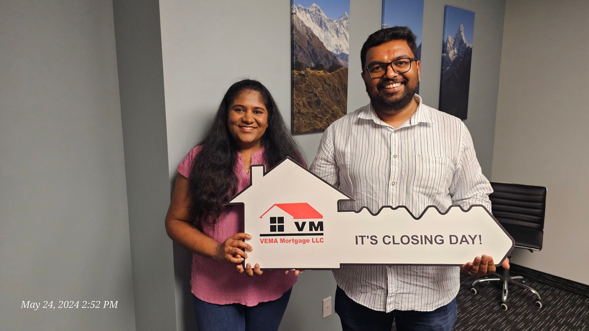 Two people holding a house-shaped sign at an event, smiling beside wall art.