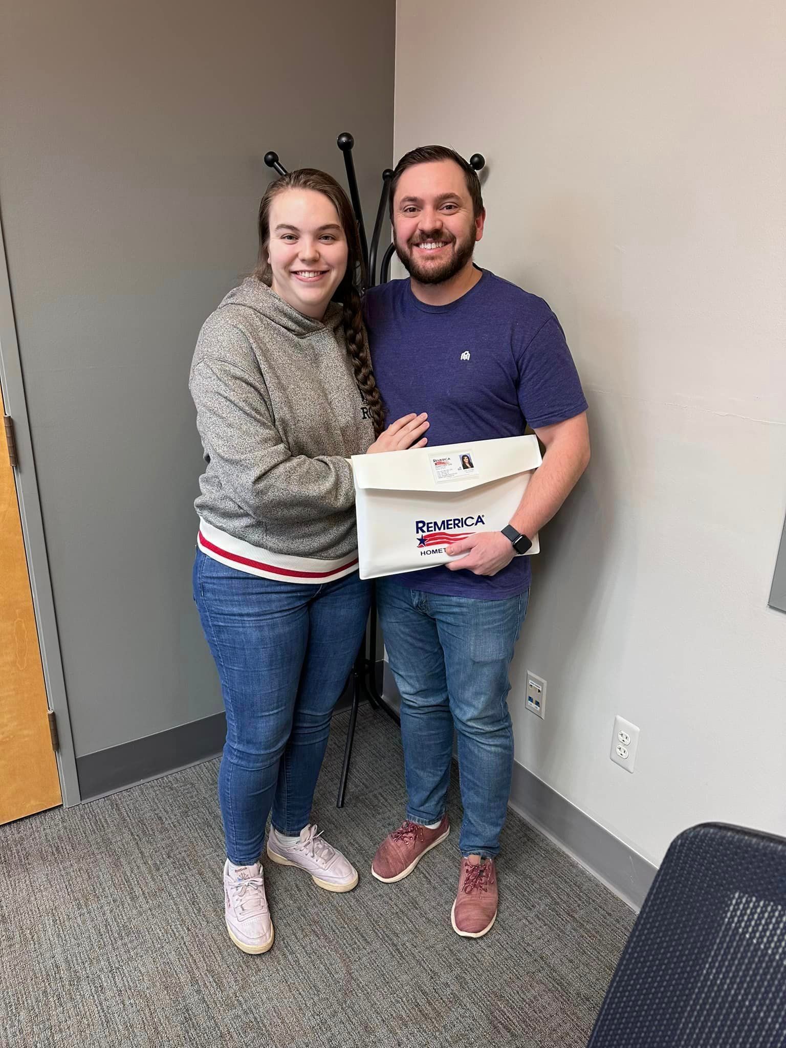 Two people smiling in a hallway, holding a small white box together.