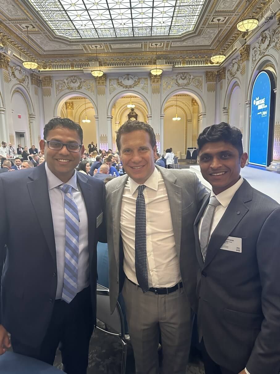 Three men in suits smiling in an ornate ballroom with chandeliers and arched mirrors.