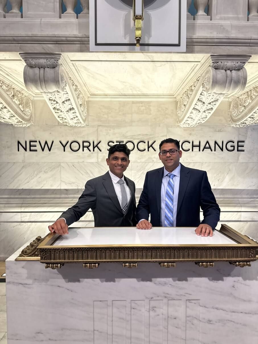 Two men in suits posing behind the New York Stock Exchange trading desk.