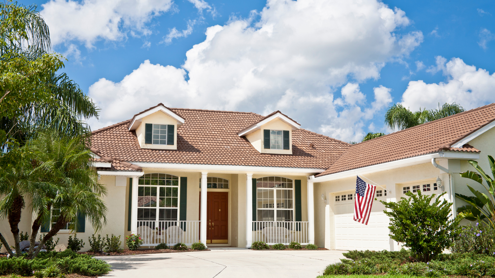 Single-story suburban house with white exterior, red tile roof, and American flag under a blue sky