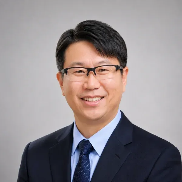 Smiling man in glasses wearing a navy suit, light blue shirt, and dark tie on a gray background