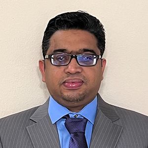 Man in a gray suit and blue shirt wearing glasses, standing against a plain wall.