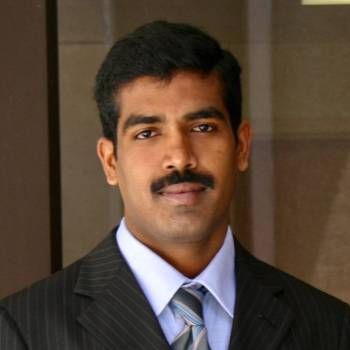 Man in a dark suit and striped tie, standing indoors with a neutral expression.