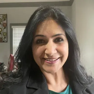 Smiling woman with long dark hair in a gray blazer, standing indoors by a window.
