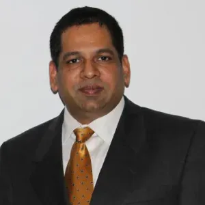 Man in a dark suit, white shirt, and orange tie against a plain light background