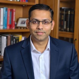 Man in a navy blazer and glasses seated before a bookshelf, facing the camera with a slight smile