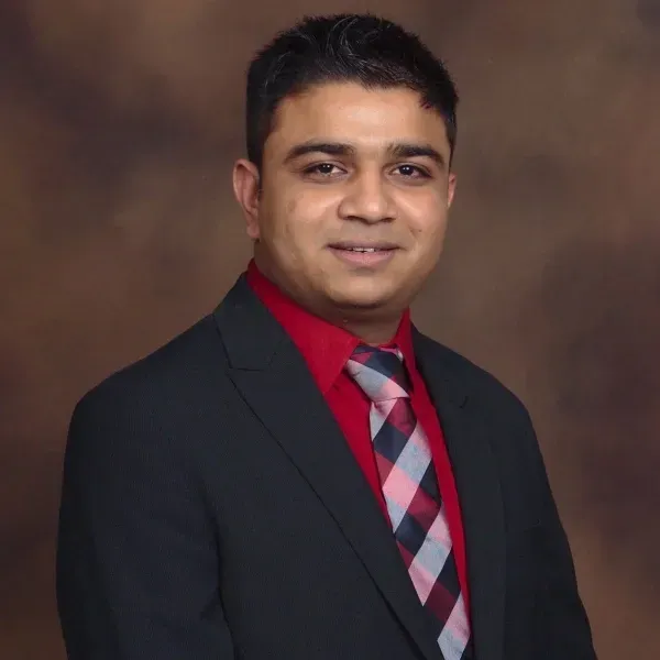 Studio portrait of a smiling person in a black suit, red shirt, and patterned tie against a brown backdrop