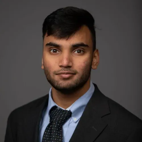 Studio headshot of a man in a dark suit, blue shirt, and patterned tie against a gray background