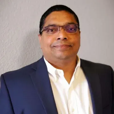 Person in a dark blue blazer and white shirt, smiling against a light gray wall.