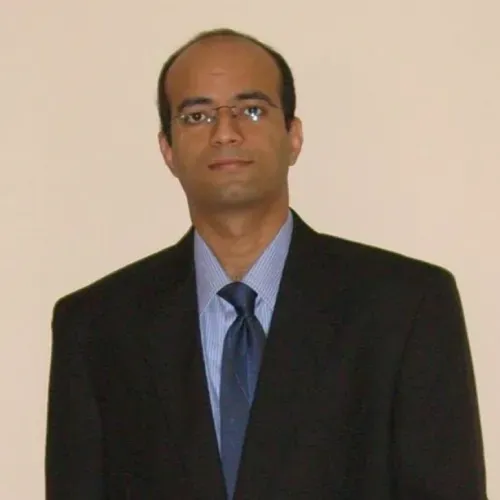 Man in a black suit and blue tie standing against a plain beige wall.