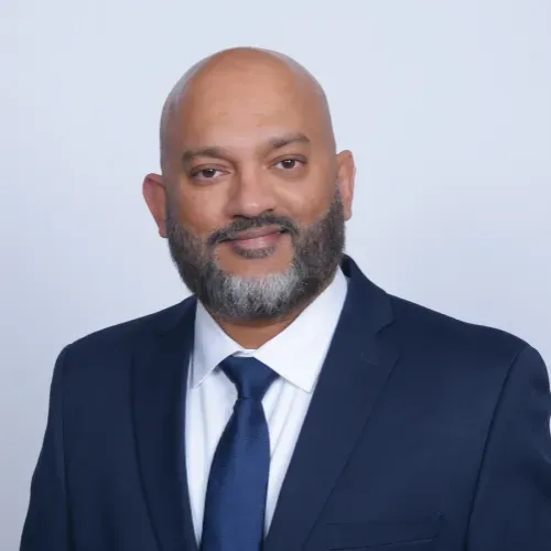 Studio portrait of a smiling bald man in a navy suit, white shirt, and blue tie on a light gray background