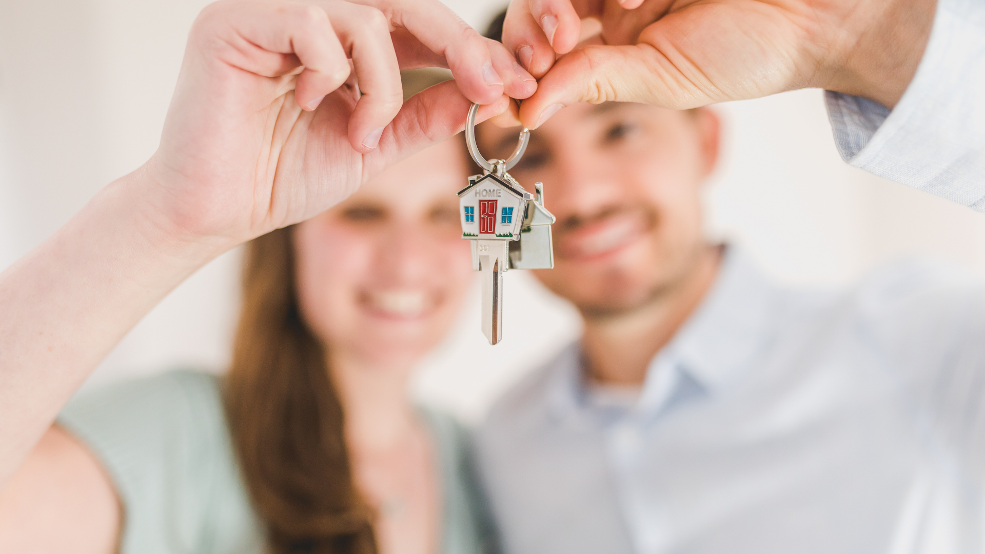 Hands holding a small house keychain in front of a smiling couple in a bright room