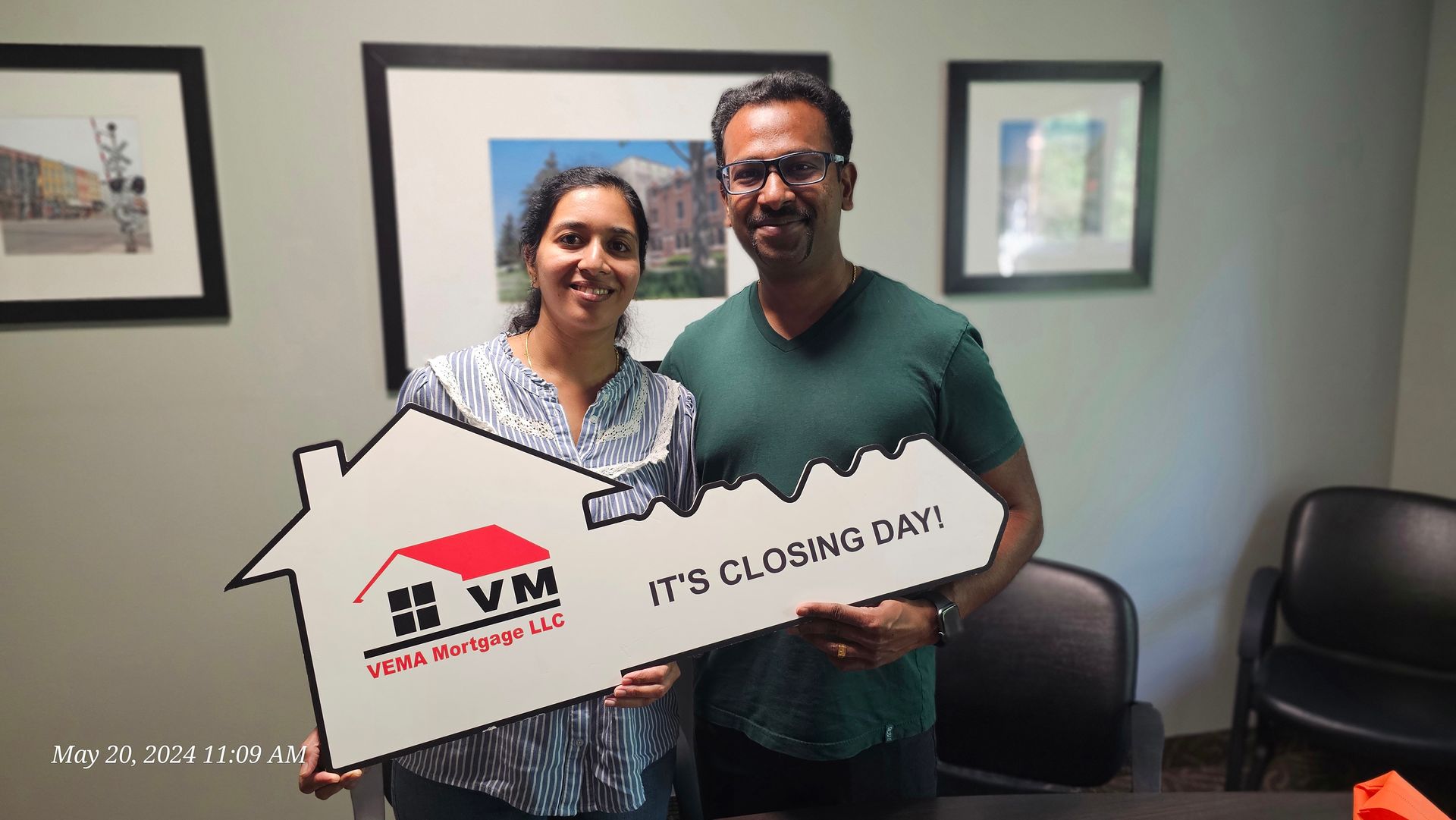 Two people holding a white house-shaped sign that says “IT’S CLOSING DAY!” in an office.