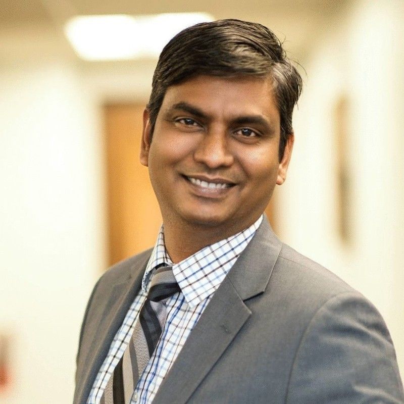 Smiling man in a gray suit and tie, standing indoors with a blurred office background