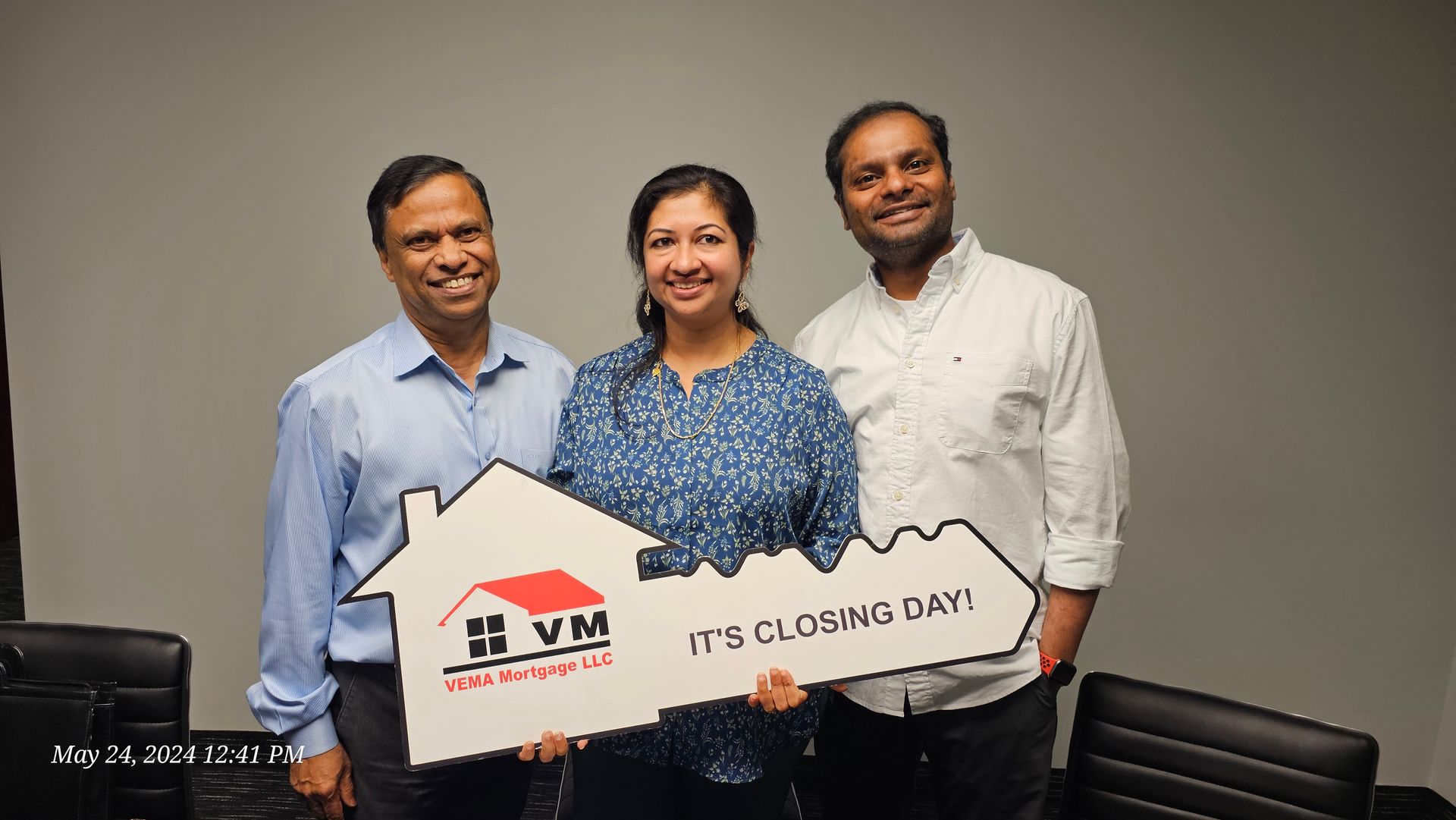Three people posing indoors with a large cardboard house cutout and a logo sign.