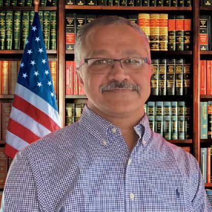 Smiling man in a checkered shirt standing before bookshelves and a U.S. flag