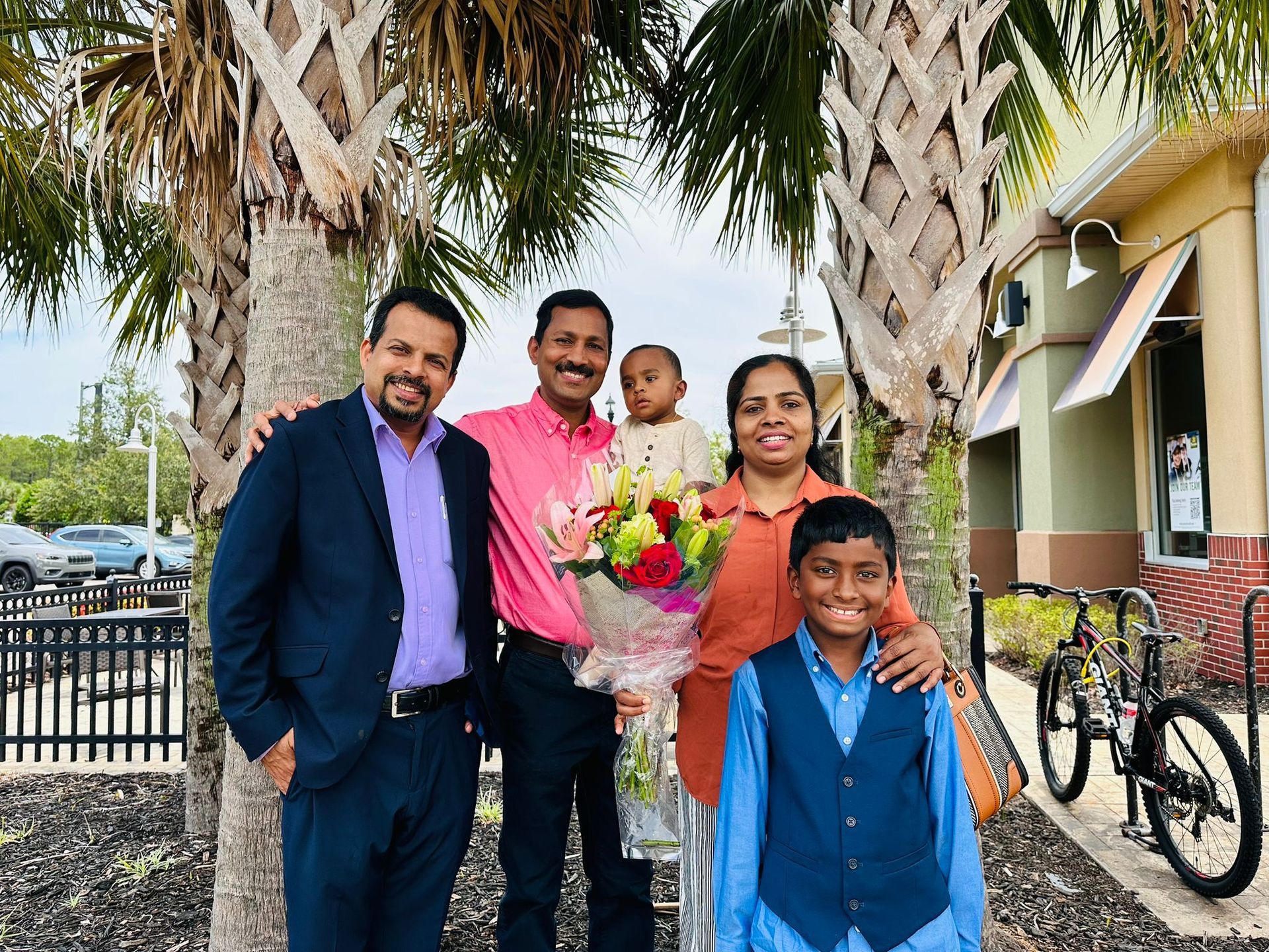 Family posing outdoors by palm trees, with a baby in a bouquet and a boy in a blue vest in front.
