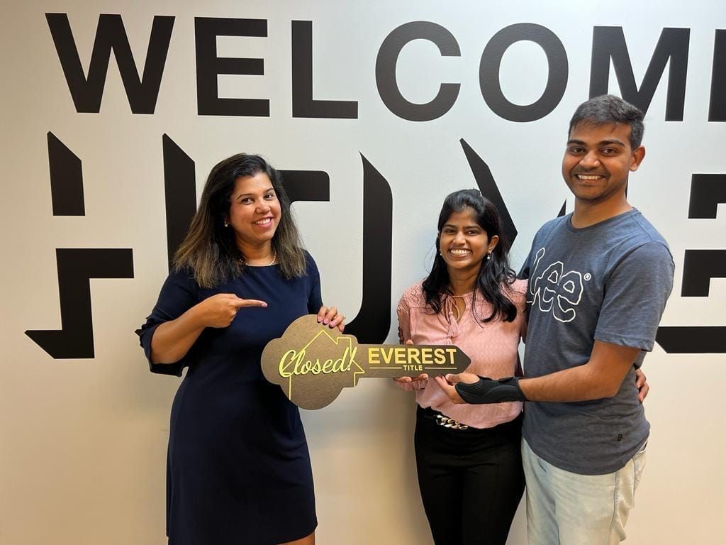 Three people holding an EVEREST sign in front of a wall with large “WELCOME” letters.