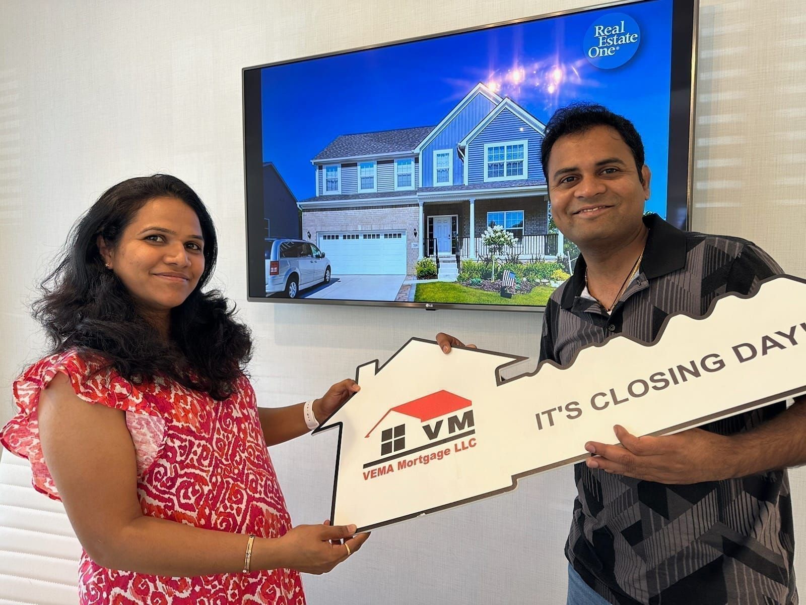 Two people holding a house-shaped sign reading “It’s closing day” in front of a home display