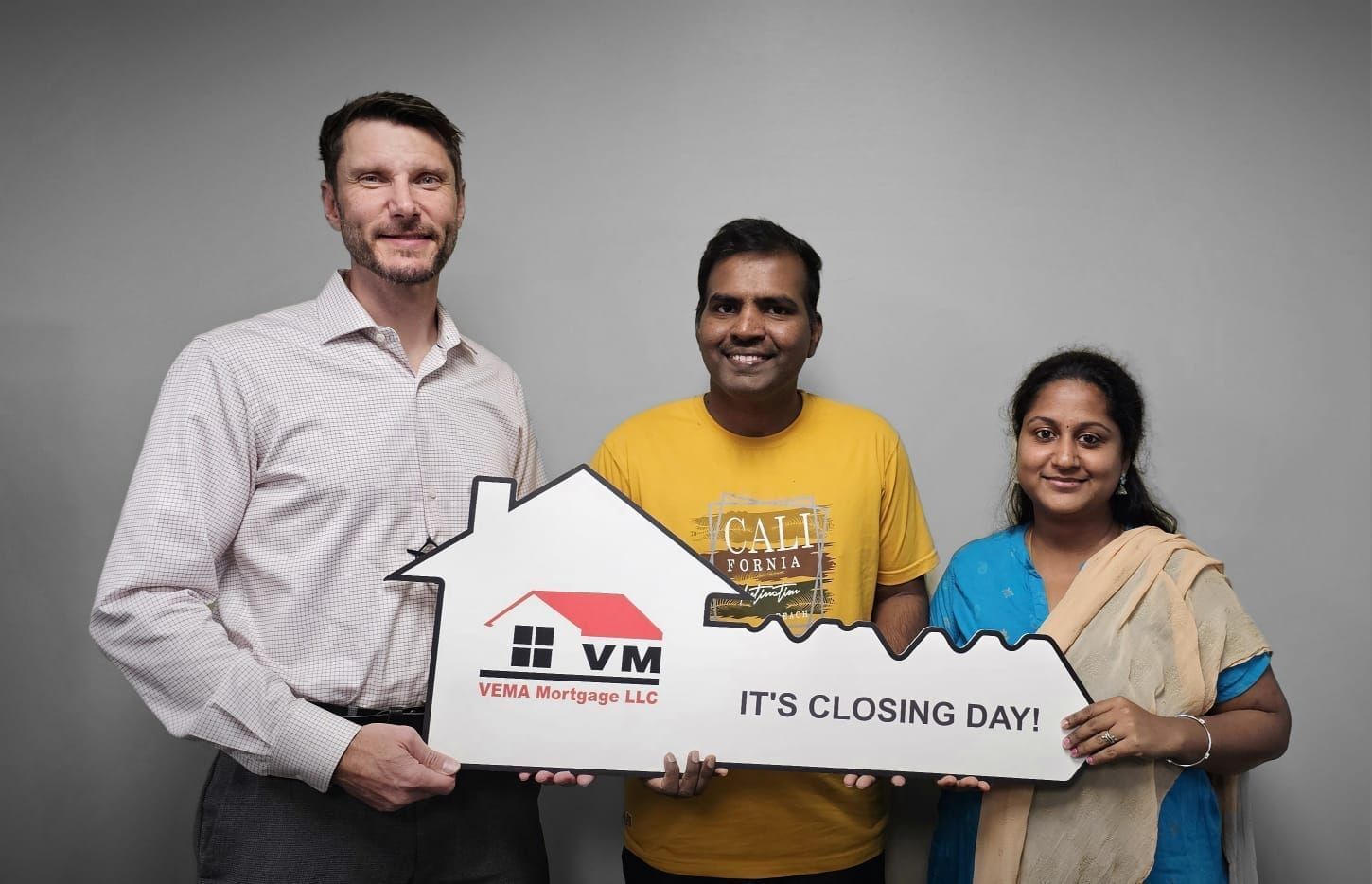 Three people holding a house-shaped sign that reads “IT’S CLOSING DAY” against a gray background