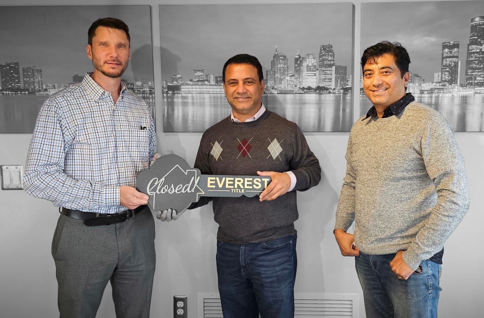 Three men stand indoors holding a branded sign in front of a city skyline backdrop.