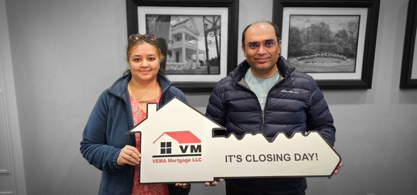 Two people holding a house-shaped sign that says “It’s closing day” in a framed office setting