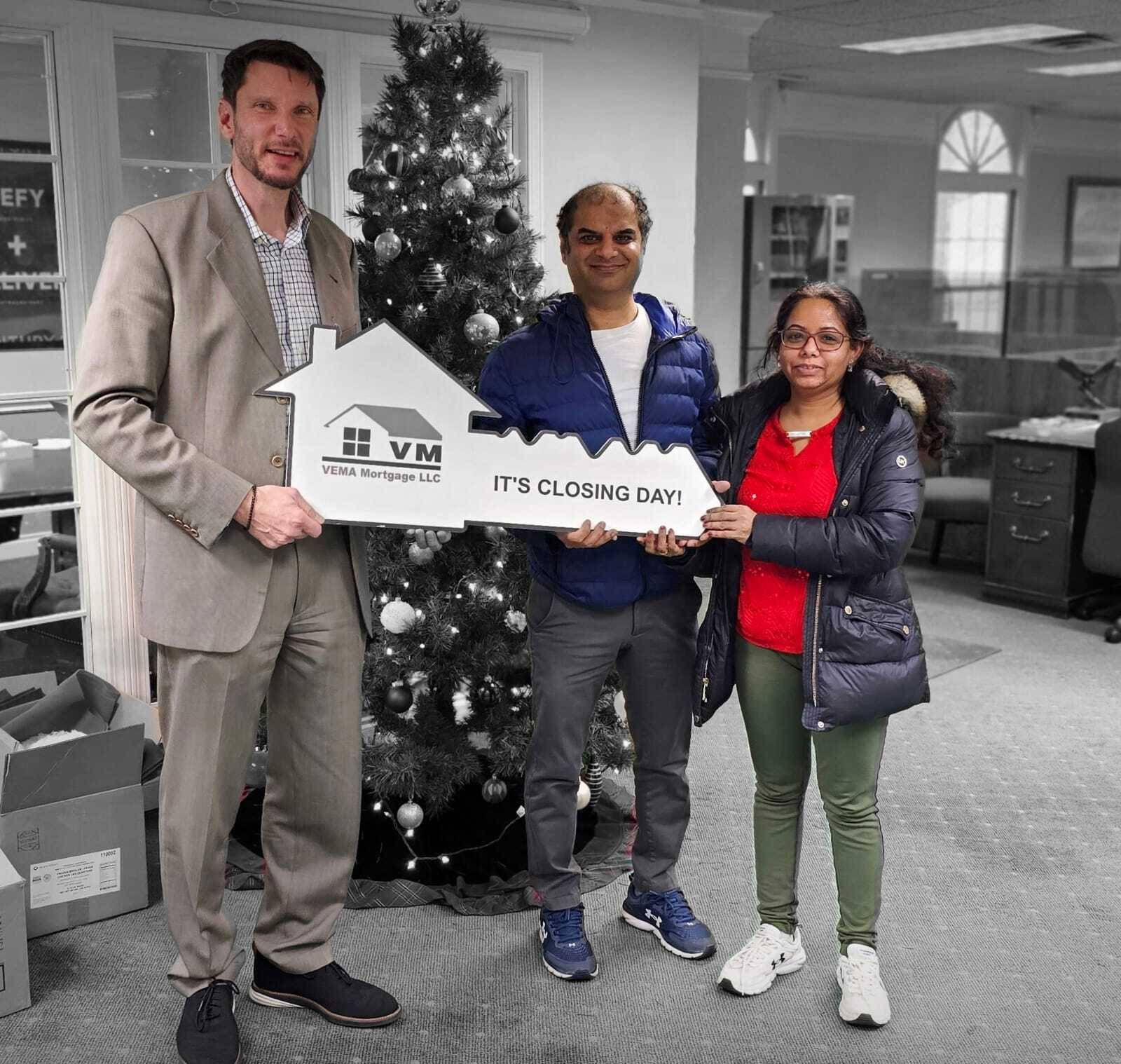Three people posing indoors beside a Christmas tree, holding a house-shaped sign.