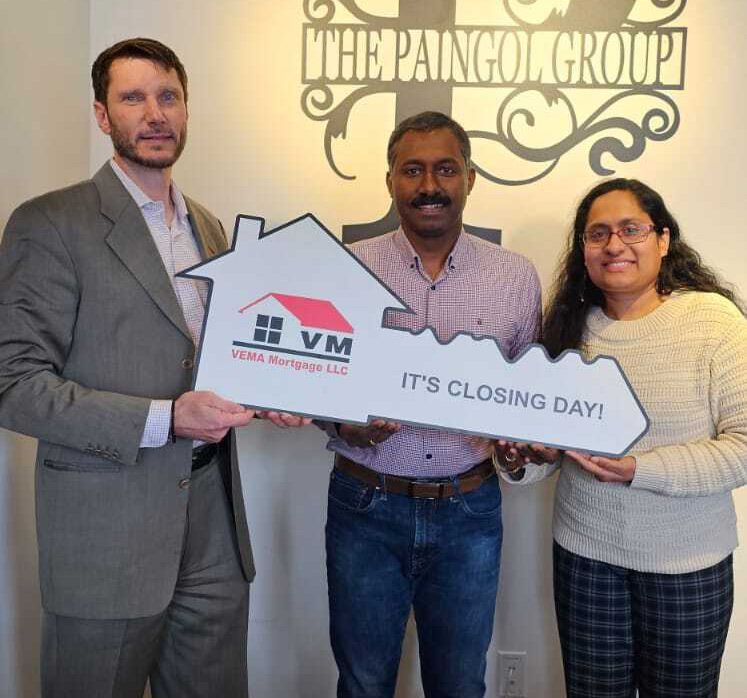 Three people holding a giant house-key prop that says “It’s Closing Day” in an office setting