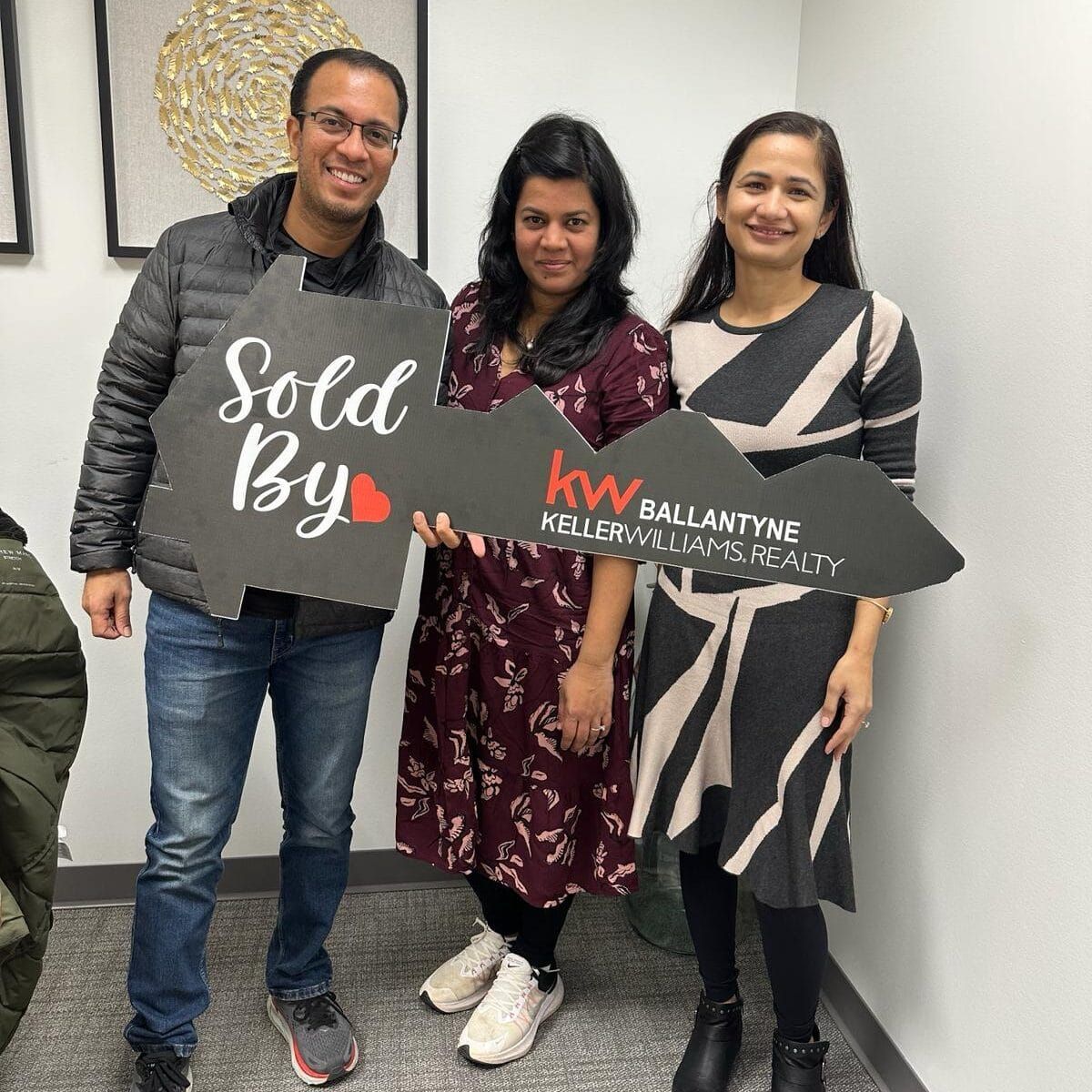 Three people posing indoors holding a large “Sold By” key-shaped sign for KW Ballantyne.