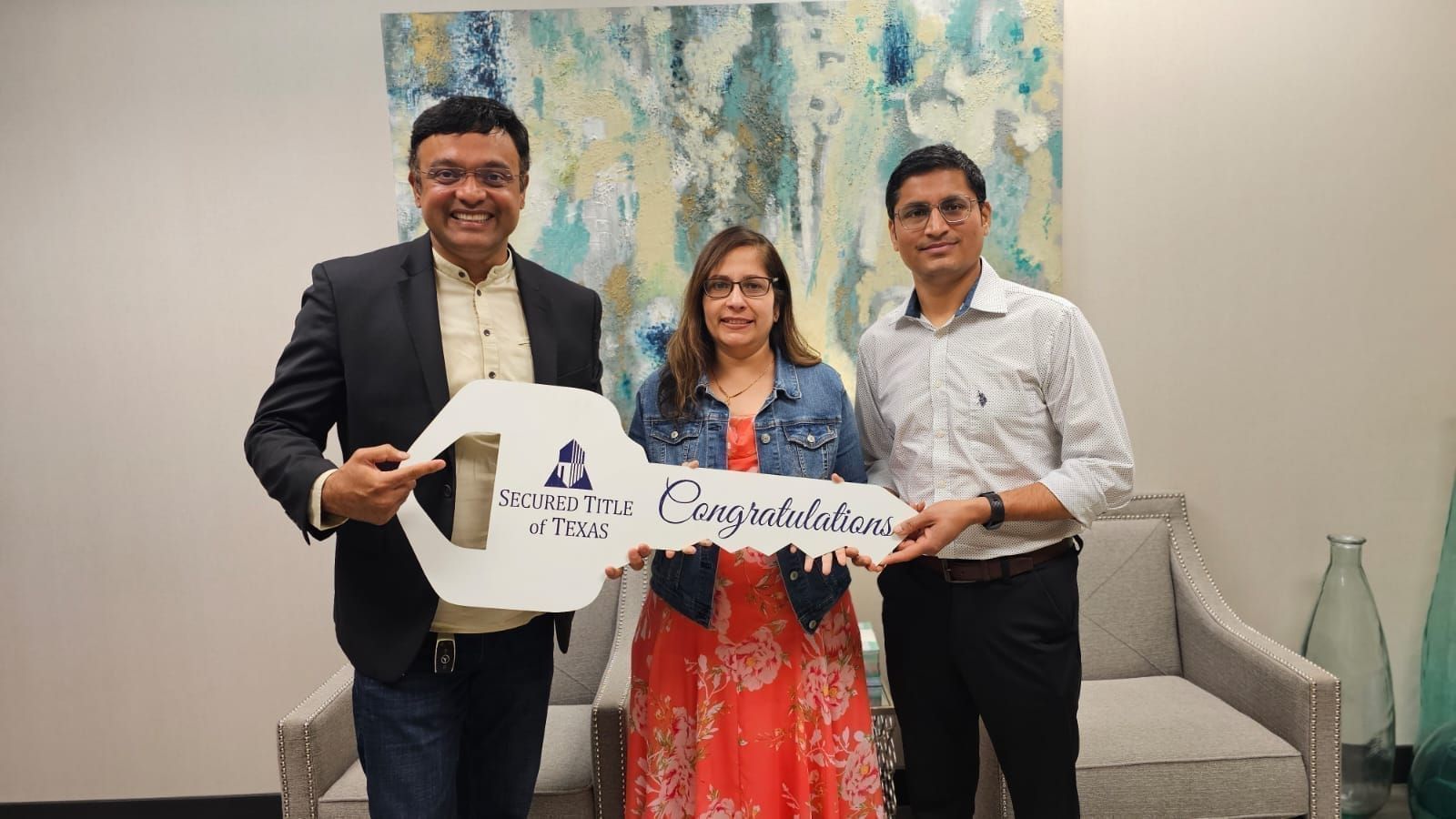 Three people hold a white banner in a meeting room, smiling in front of abstract wall art.