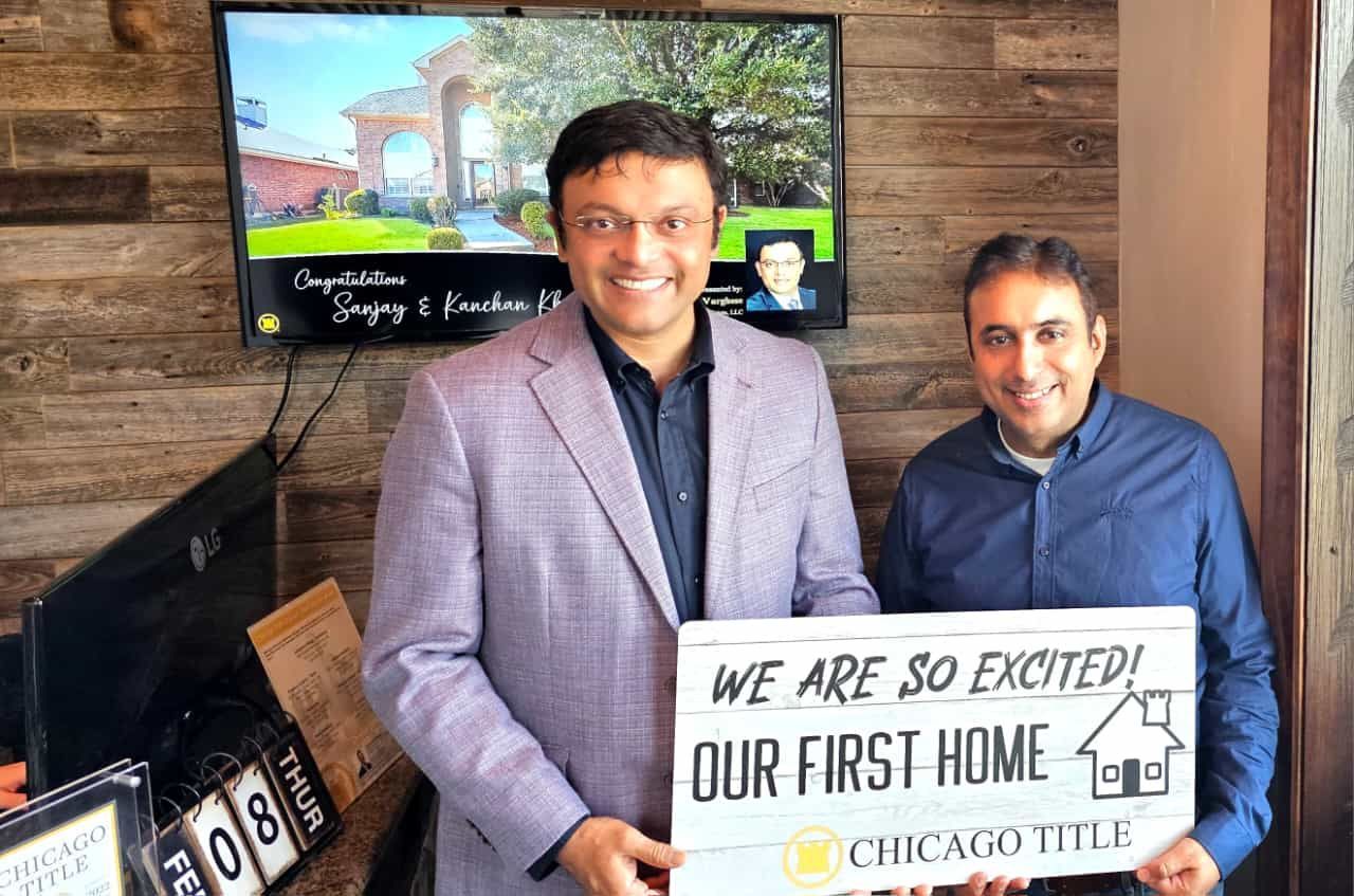 Two men indoors holding a “We are so excited! Our first home” sign in an office setting