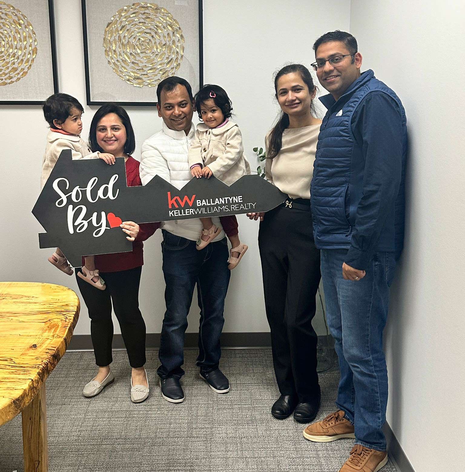 Family holding a “Sold By” real estate sign in an office, smiling beside a realtor team.