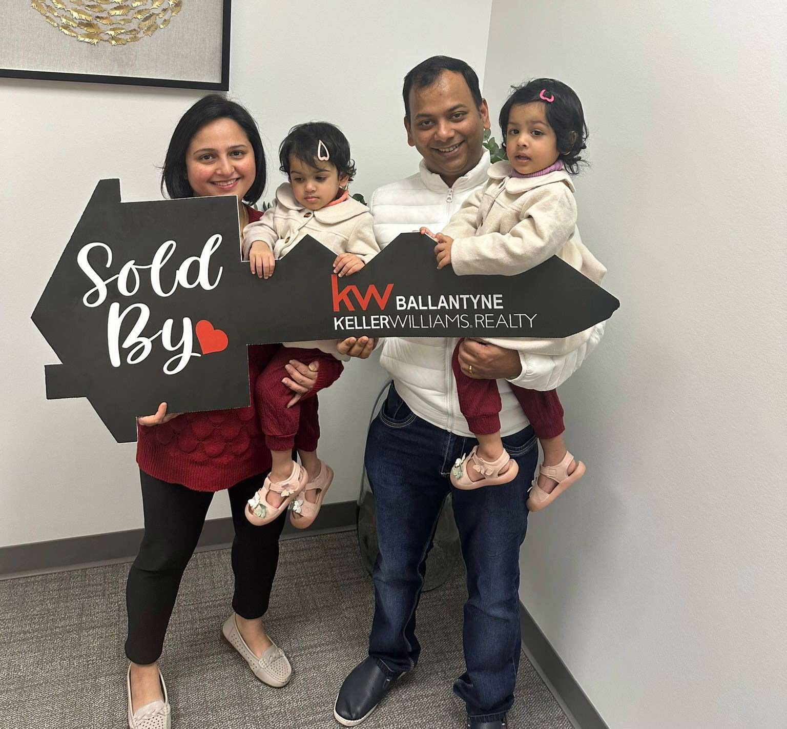Family posing indoors with two sold signs, smiling and holding a baby and toddler