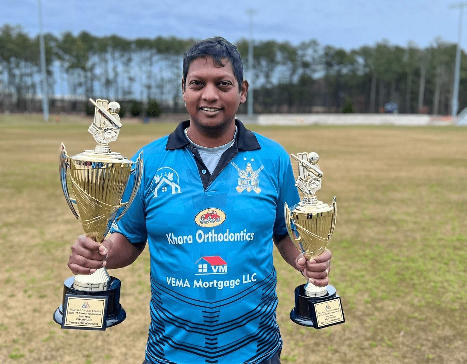 Smiling person in blue sports jersey holding two gold trophies on a cricket field