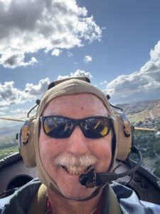 Smiling pilot wearing sunglasses and headset in a cockpit under a partly cloudy sky