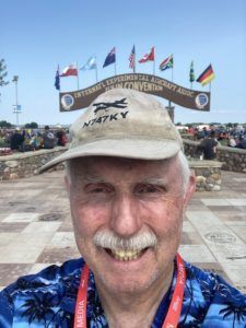 Smiling man in a tan cap stands before a flag-lined memorial entrance under cloudy skies
