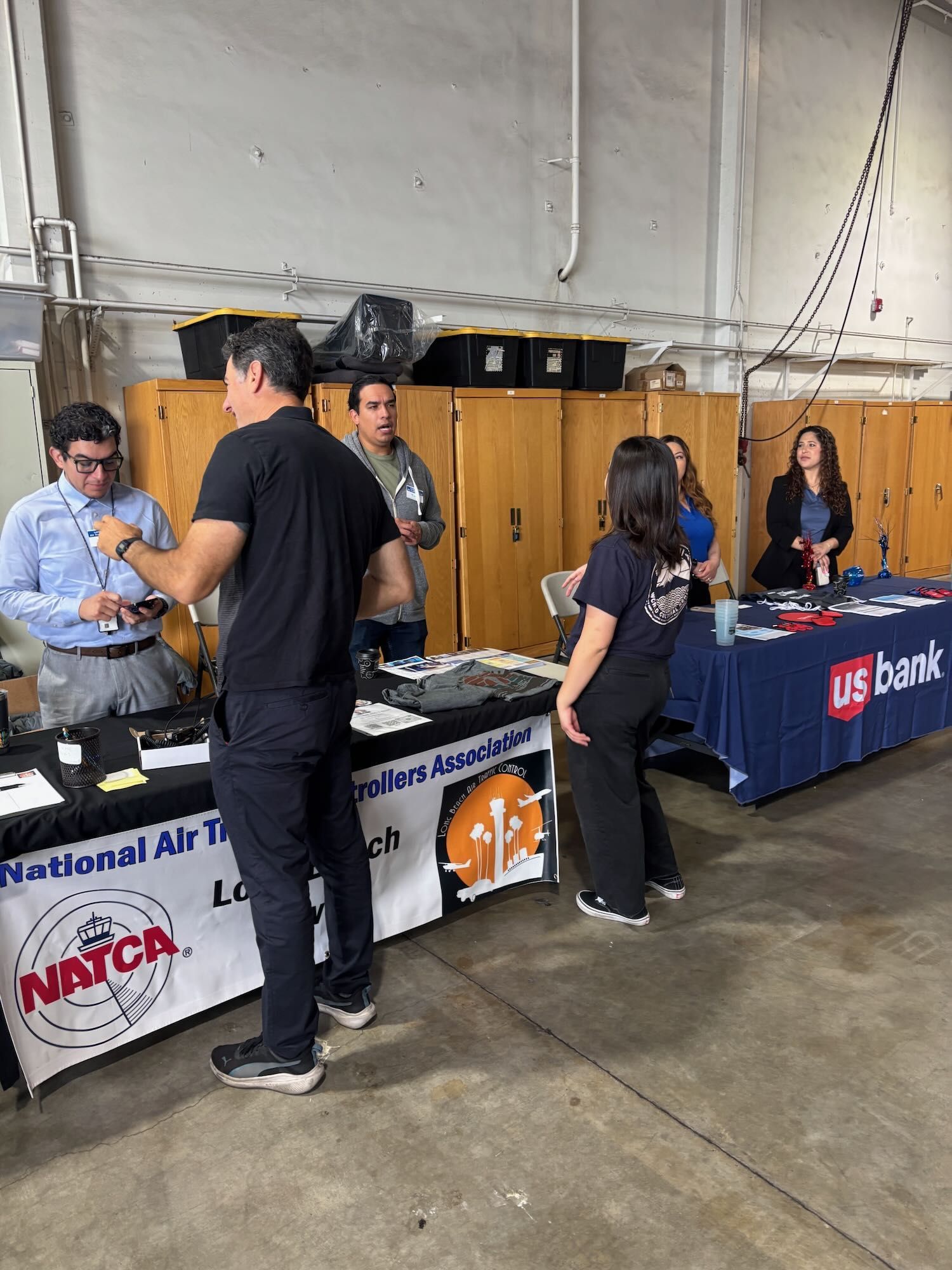 People at a bank booth in a gymnasium, with tables, banners, and flyers during an event.