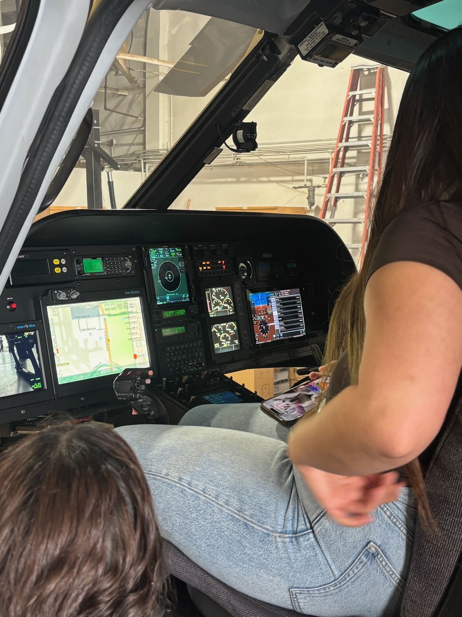 Two people seated in a helicopter cockpit, with illuminated controls and a ladder visible outside.