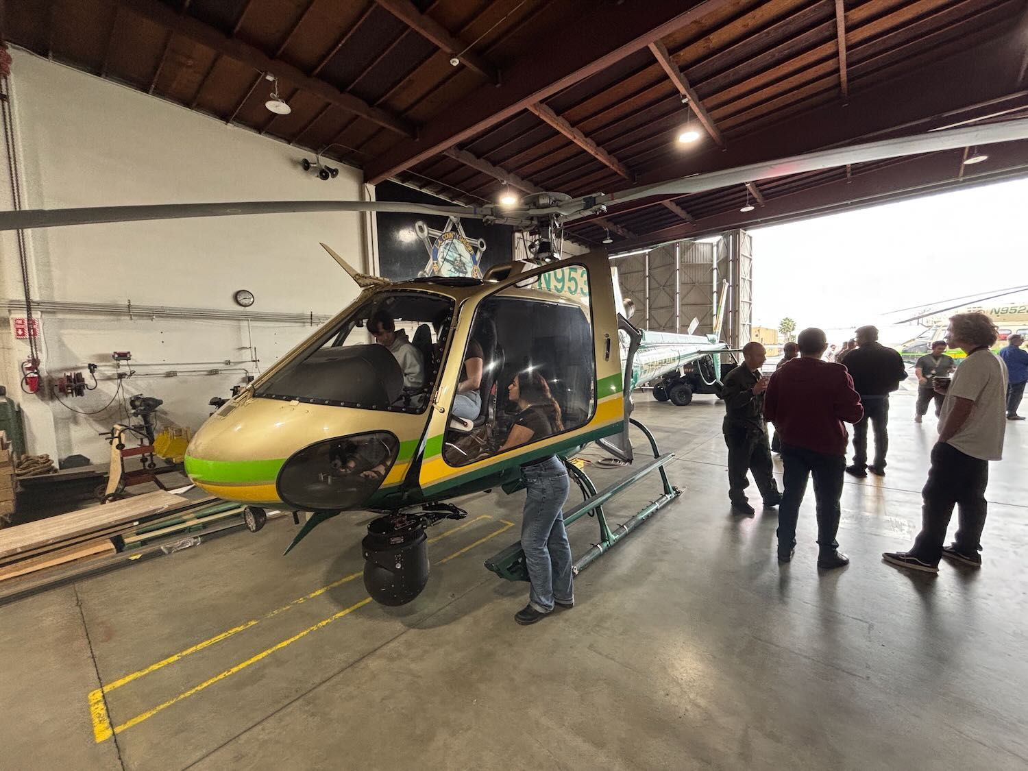 People gathered around a yellow helicopter inside an industrial hangar.