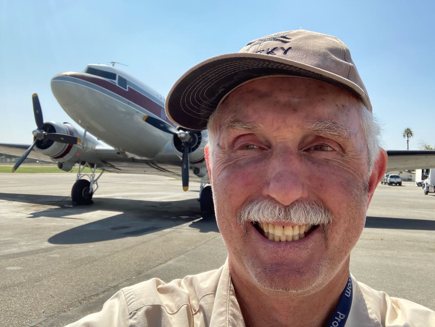 Smiling man in a cap taking a selfie at an airport with a small airplane behind him
