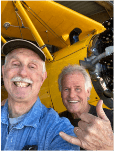 Two smiling men posing in front of a yellow aircraft, one making a shaka hand gesture.