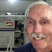Smiling man in foreground beside industrial equipment and a lit display in a workshop