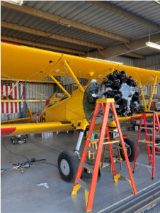 Yellow airplane in a hangar with two ladders and an exposed engine being repaired