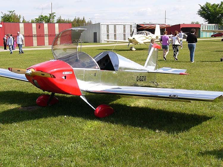 Small red-and-silver model airplane on grass at an outdoor airfield with people in the background
