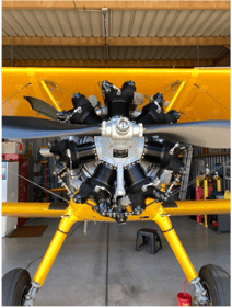 Front view of a yellow aircraft with a black propeller and radial engine inside a hangar