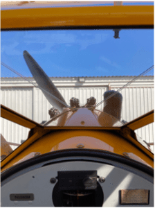 View from inside a yellow biplane cockpit, looking past the propeller toward blue sky and a hangar.
